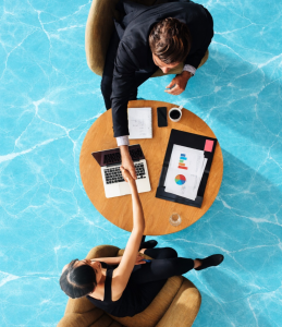 Deux professionnels échangent une poignée de main au-dessus d’une table ronde avec ordinateur portable et graphiques financiers, symbolisant la réussite financière et la collaboration, sur fond bleu clair